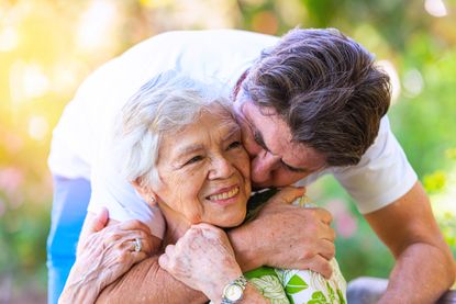 Mature man hugging his senior smiling mother while visiting her at a nursing home.