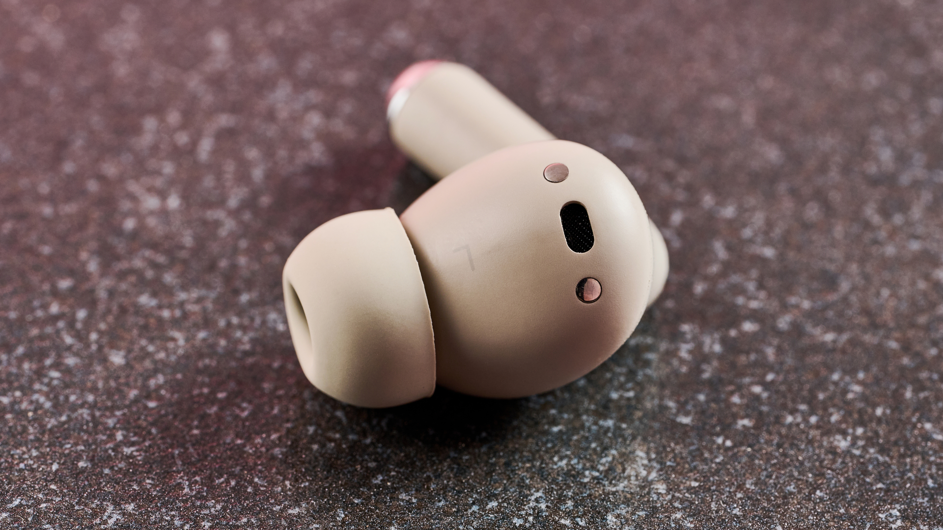 A close up view of one of the Motorola Moto Buds Plus earbuds. You can easily see the silicone tip and the microphone at the top of the ear piece. It is siting on a dark work surface.