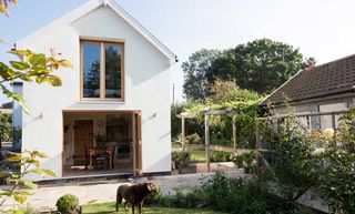 a white house with wooden window and door frames, with an eclectic kitchen extension inside full of antique pieces, and a lush garden in the foreground