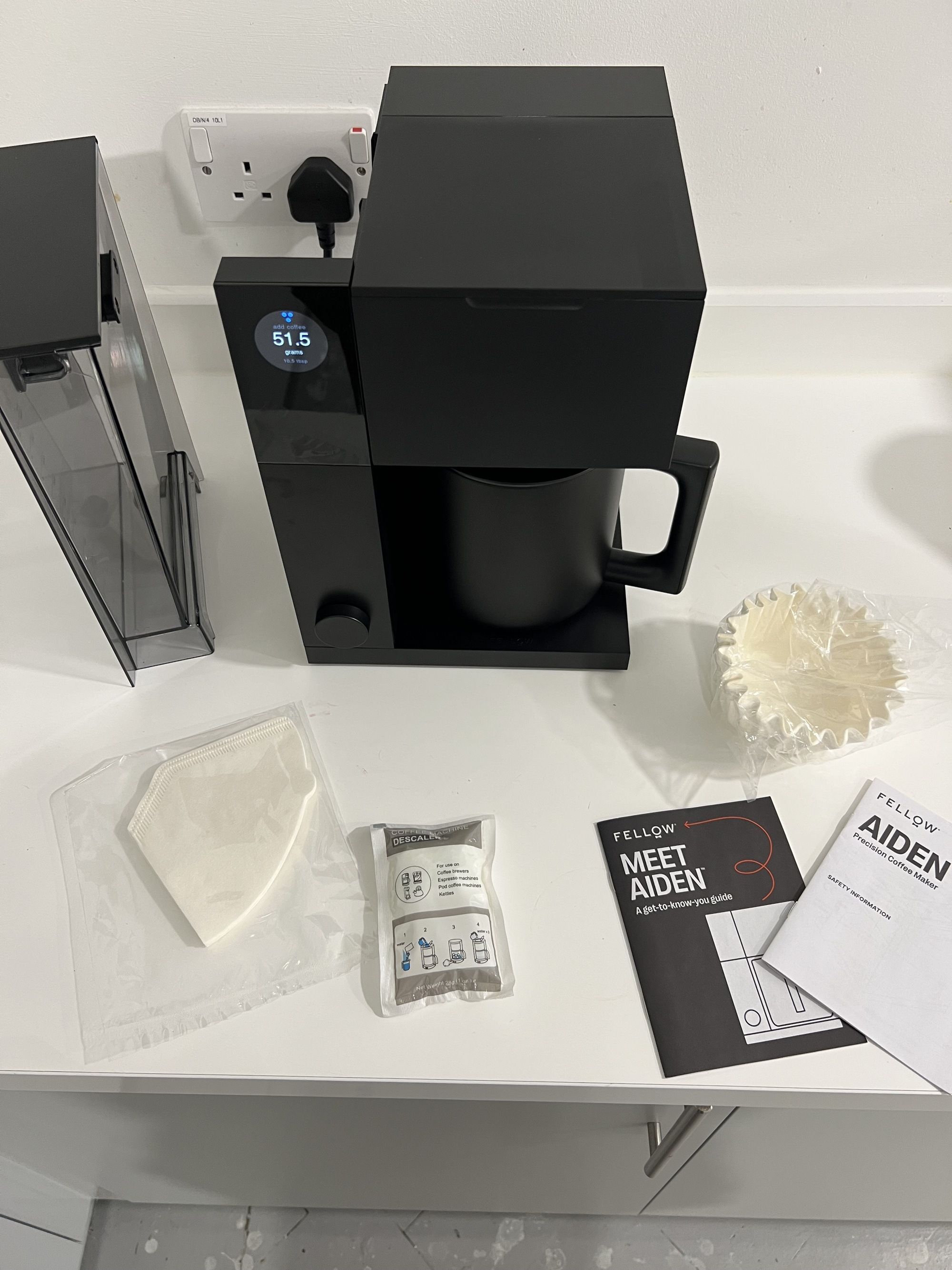 Image of the Fellow Aiden Precision Coffee machine in black on a white countertop with instruction manuals and coffee filter around it.