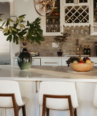A close shot of a white kitchen island with a green vase of stems and a wooden fruit bowl on it, with three chairs in front of it and whitte cabinets and a silver fridge behind it