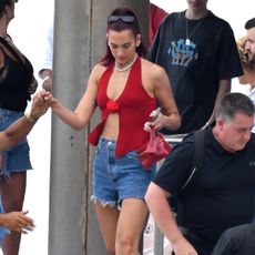 Dua Lipa wearing a red With Jean halter top with jean shorts, black sunglasses, and a red handbag.