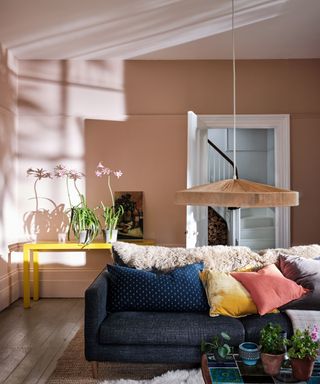 A pink living room with a blue couch, yellow side table and a low hanging wooden pendent light