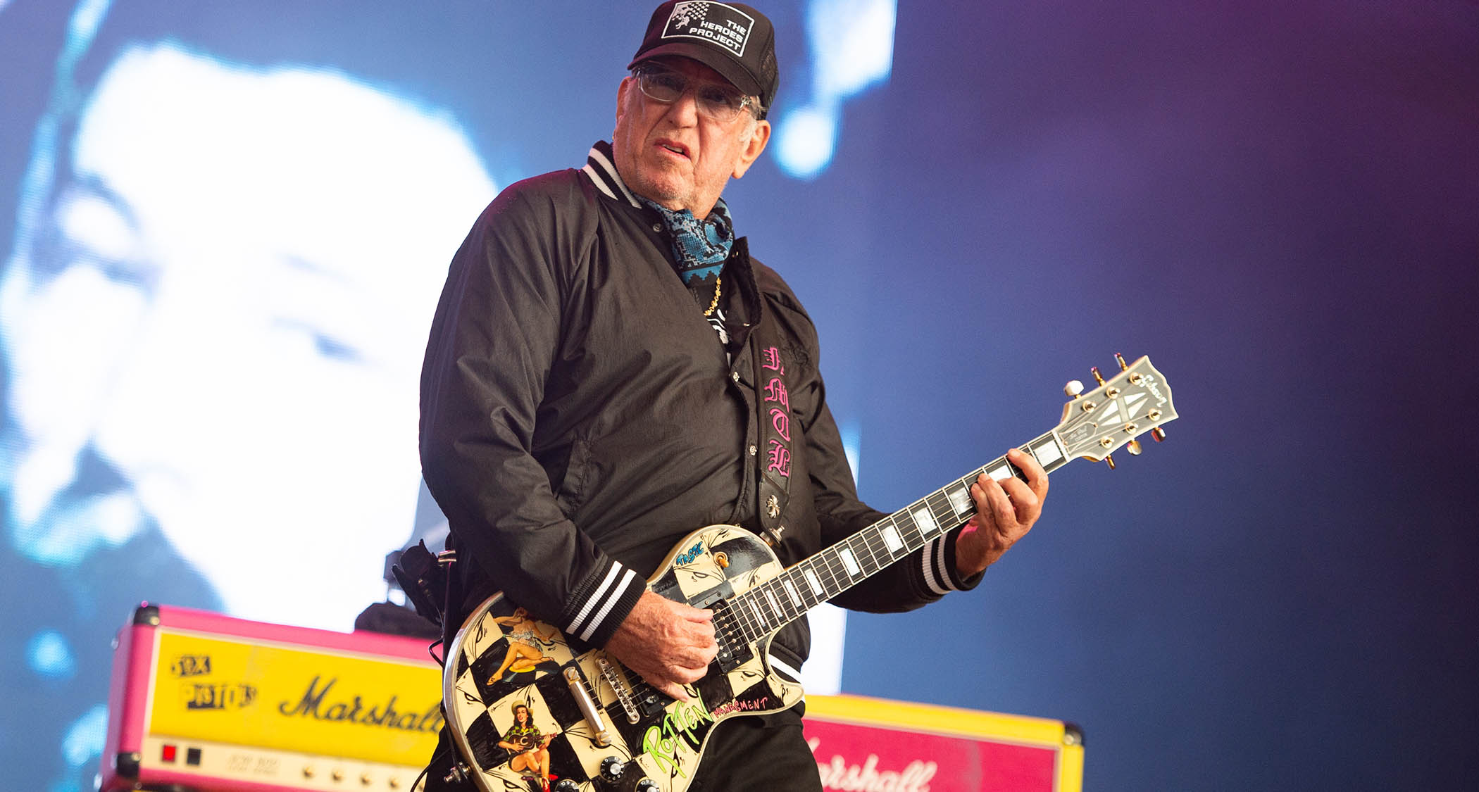 Steve Jones of the Sex Pistols wears a black ballcap and plays his Les Paul Custom as he performs at Download, day-glo Marshall amps in the background.
