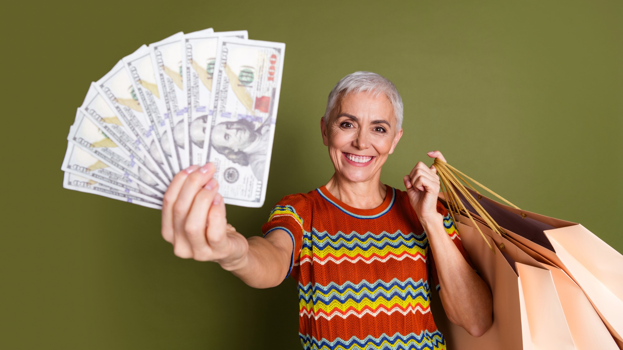 Portrait of smiling happy mature woman holding dollar bills and shopping bags on green background