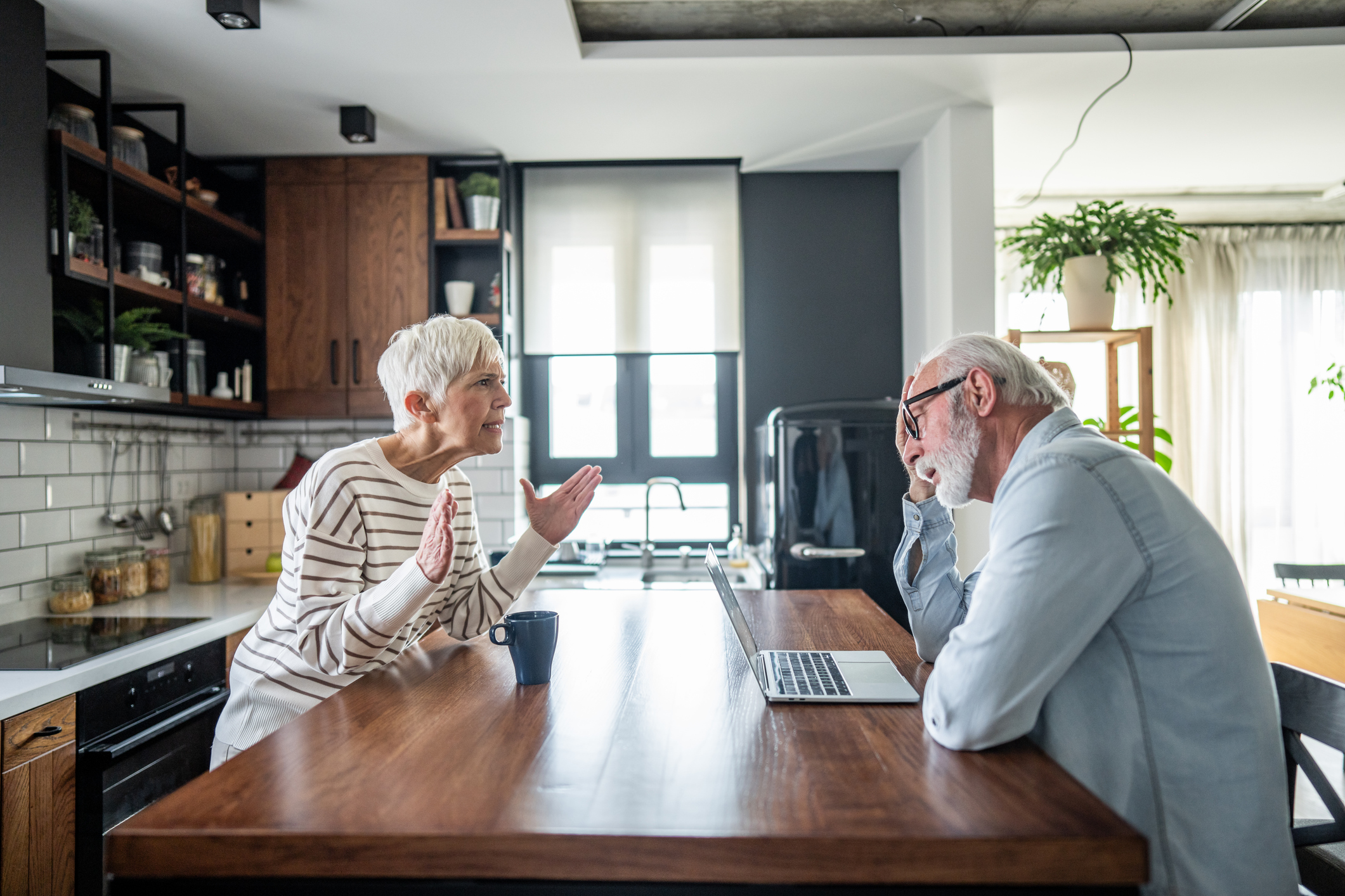 Senior couple having a disagreement while using a laptop in their modern kitchen, highlighting communication challenges and relationship dynamics in later life.