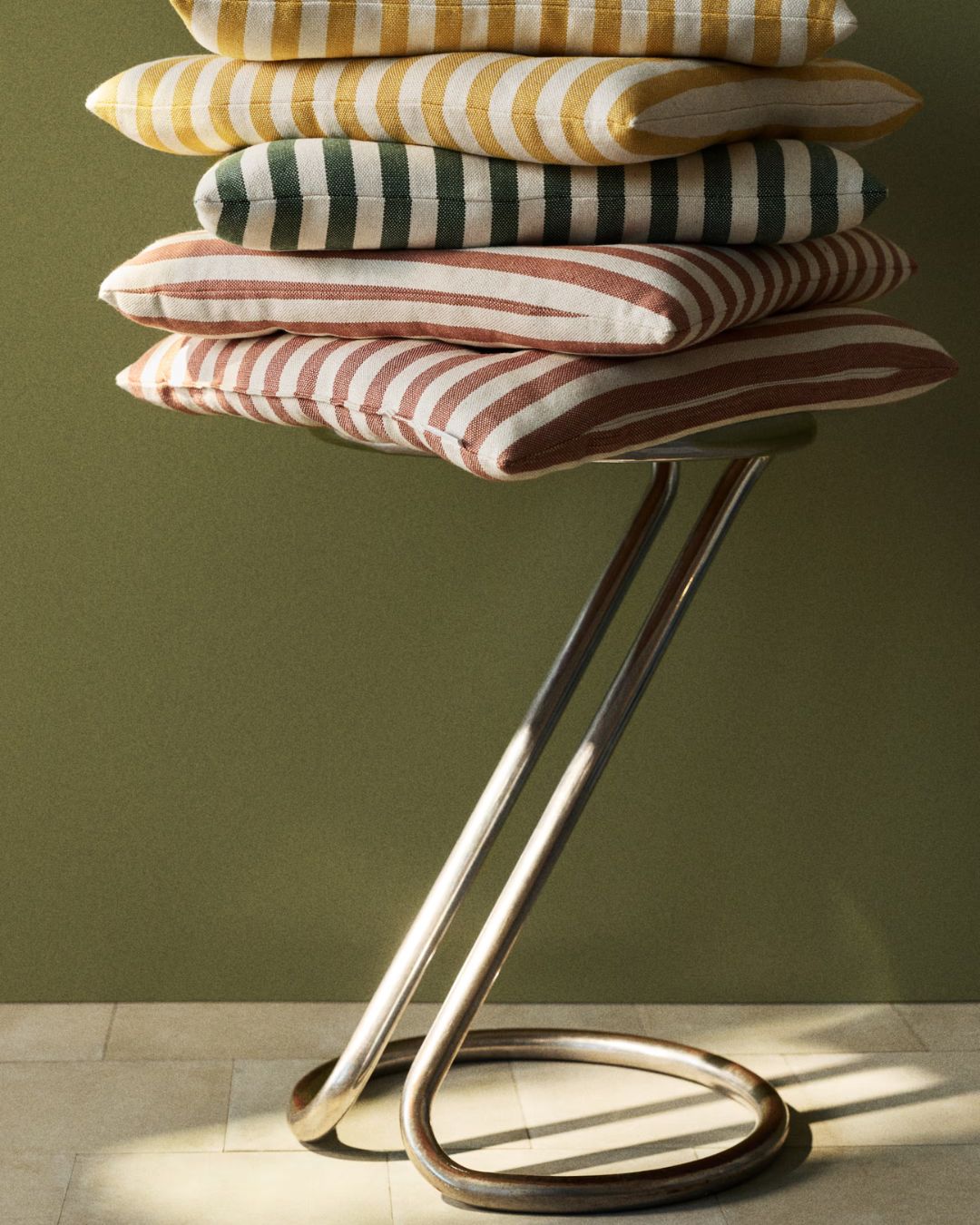 Outdoor Seat Cushions piled up on a sculptural chrome stool against an olive green background.