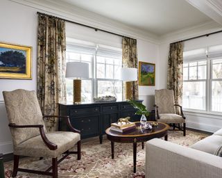 A living room with patterned curtains, a black sideboard with studded lamps and patterned dark wood armchairs