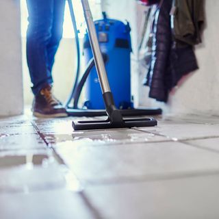 Man in jeans and brown shoes vacuuming water spill on tiles