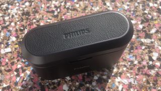 Ett svart laddningsfodral för Philips Fidelio T1 mot en spräcklig bakgrund. Laddningsfodralet är vänt uppåt med Philips-logon på locket.