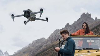 DJI Air 3 en vuelo junto a una montaña, con una persona mirando el controlador al lado de un todoterreno amarillo