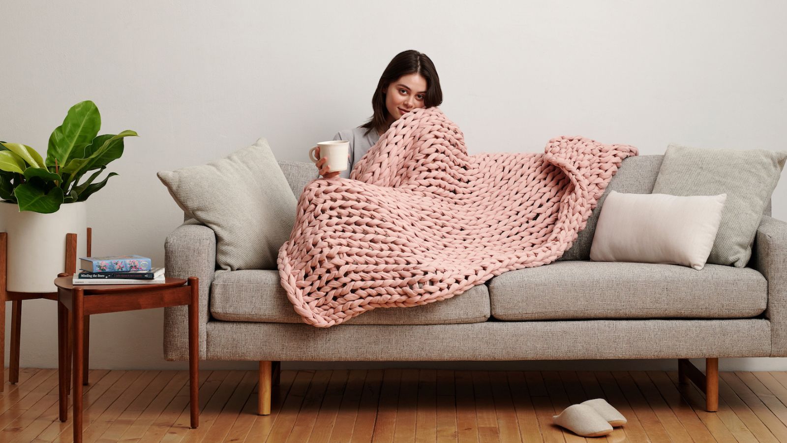 A woman lounging on a couch with a Bearaby Weighted Cotton Napper and coffee in hand.