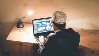 A man in a tinfoil hat hunches over a laptop