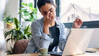 A woman looking stressed and holding the bridge of her nose while sitting at a table and using a laptop