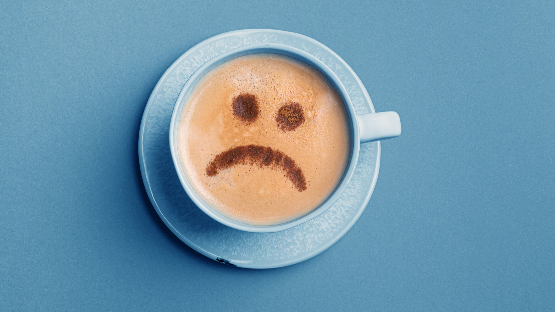 A blue cup and saucer with a brown liquid in it and a sad emoji drawn with coffee.