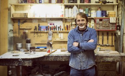 An older man poses in his workshop. He appears to be an avid carpenter or woodworker.
