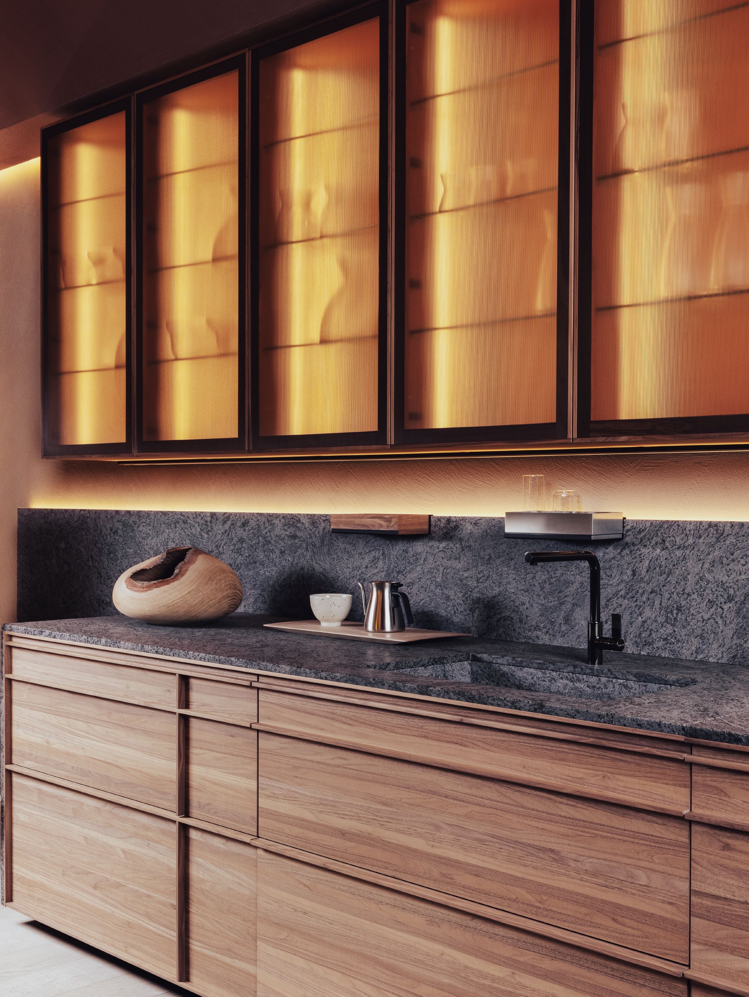 A modern kitchen featuring wooden cabinetry, a dark stone countertop, and illuminated glass-front cabinets, showcasing a minimalist design.