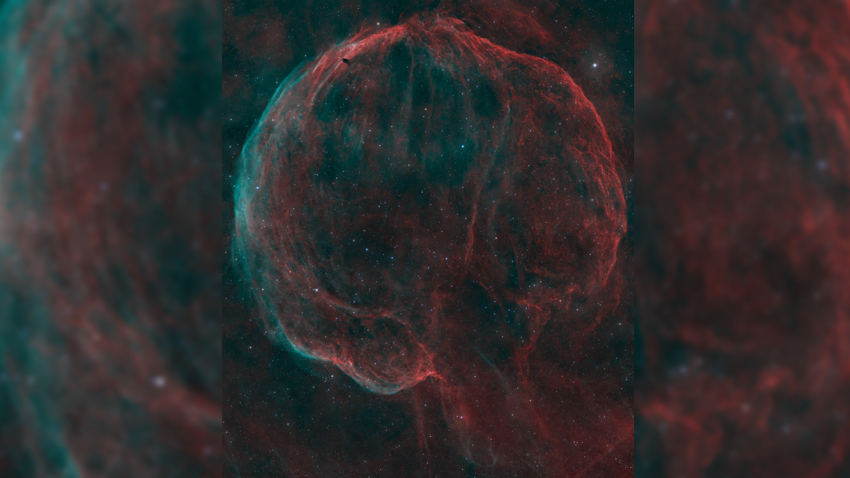 A deep space nebula scene showing a bubble-like shell of cosmic dust and gas shining red and blue against a black sky peppered with bright stars.