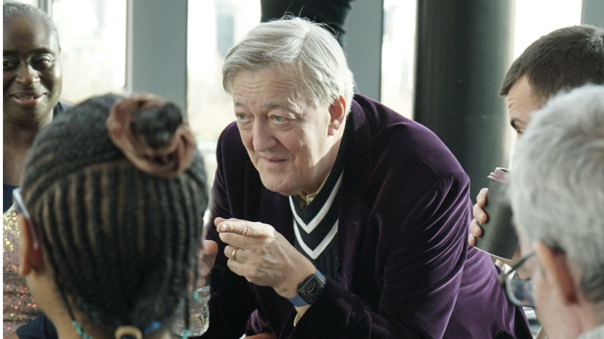 Sir Stephen wearing a purple jumper, leans into a group of interviewers in "The Assembly" season 2