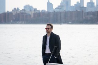 Matt Murdock (Charlie Cox) stands on the waterfront, with a view of the New York City skyline behind him. He is wearing red-tinted glasses, and his walking cane is resting against a bollard in front of him. He is wearing a suit with no tie and his top button undone, with a long navy overcoat over the top. He has his hands in his trouser pockets.