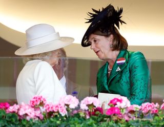 Queen Camilla wears a white suit and hat while standing with one of her companions, Lady Katharine Brooke, who wears a green outfit with a black hat at Ascot