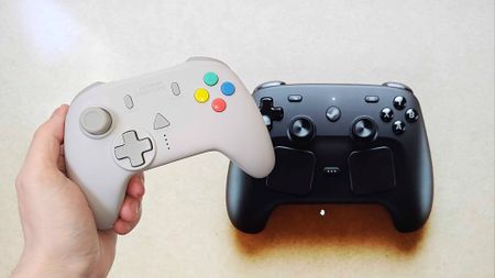 Hand holding Retro Fighters StrikerDC Dreamcast controller next to Valve Steam Controller on screen in backdrop.