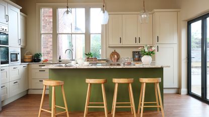 Green kitchen island with marble-effect surface, wooden bar stools and off white coloured units