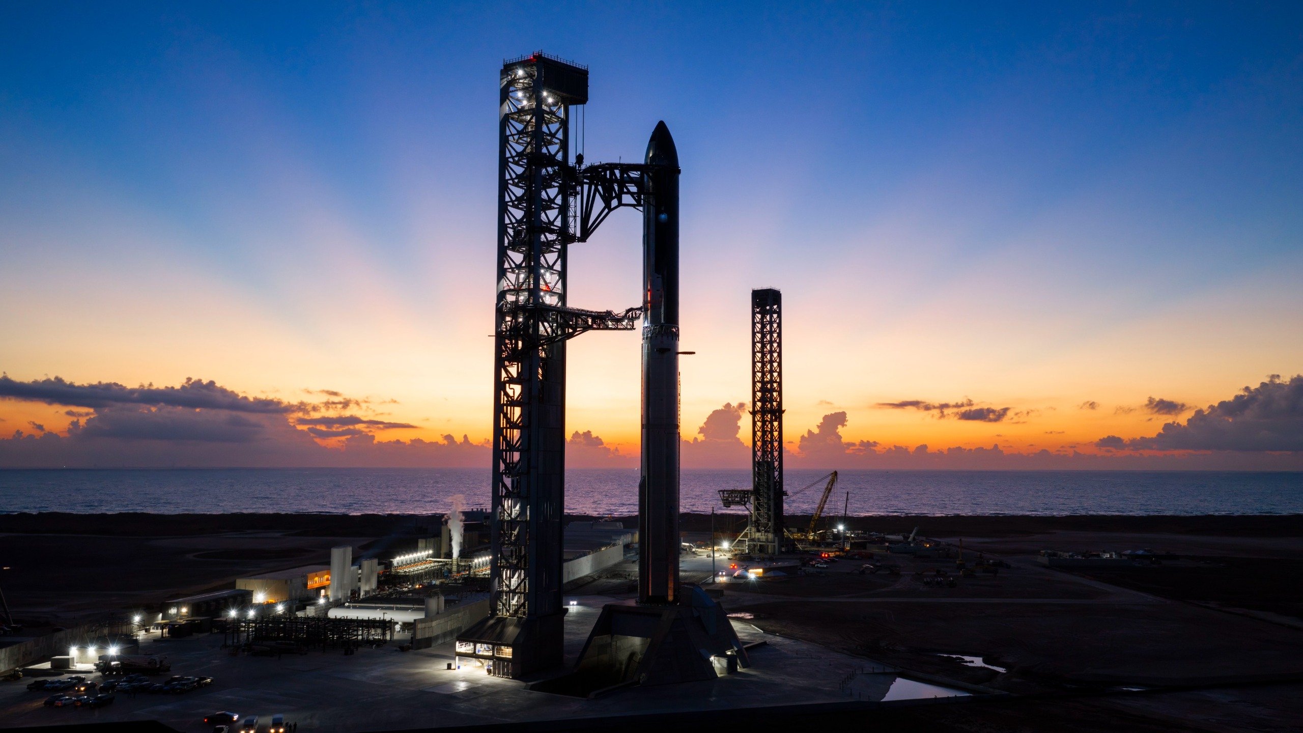 A towering SpaceX Starship rocket stands at Starbase's Pad 2, silhouetted against a dark blue sky and yellow rays from a rising sun.