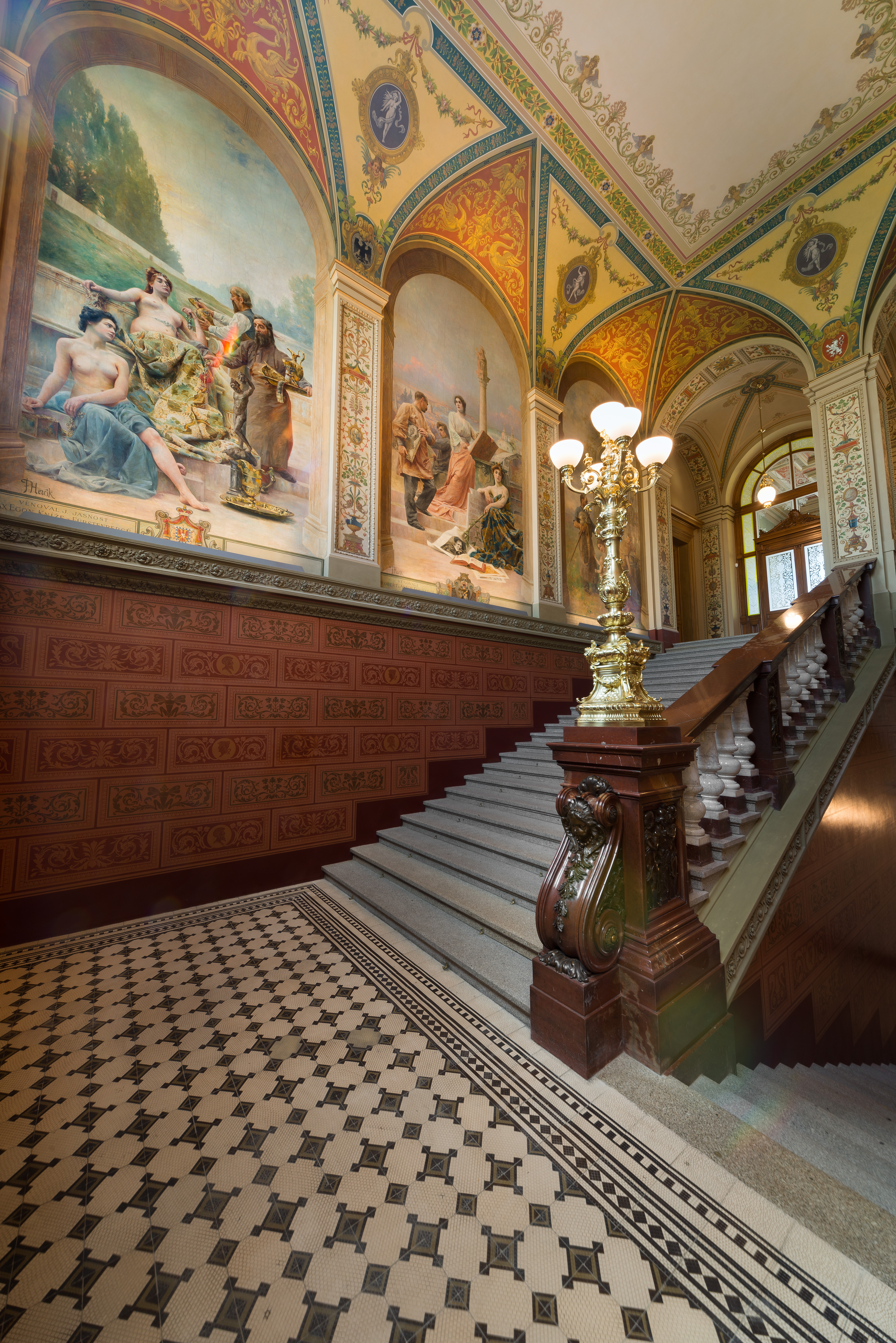 A staircase of a historical, palatial building features patterned tiled floors in black and white, half-tiled walls in terracotta, a series of colorful frescoes depicting women at leisure, gold-clad lamp posts, and wooden handles.