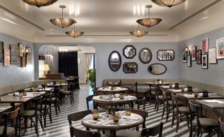 A restaurant in the Redchurch Townhouse hotel. The area is covered with round wooden tables and chairs. The walls are painted in gray color and have a lot of mirrors and pictures in frames hanging from it.