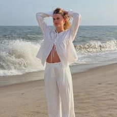 A woman at the beach wearing white linen trousers and a front tie linen long-sleeve top. 