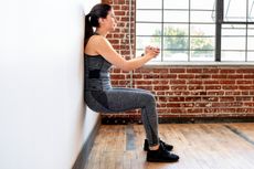 A woman performs a wall sit exercise in a room with exposed brickwork and wooden flooring. Her back is against the wall, knees bent, hands clasped in front of her.