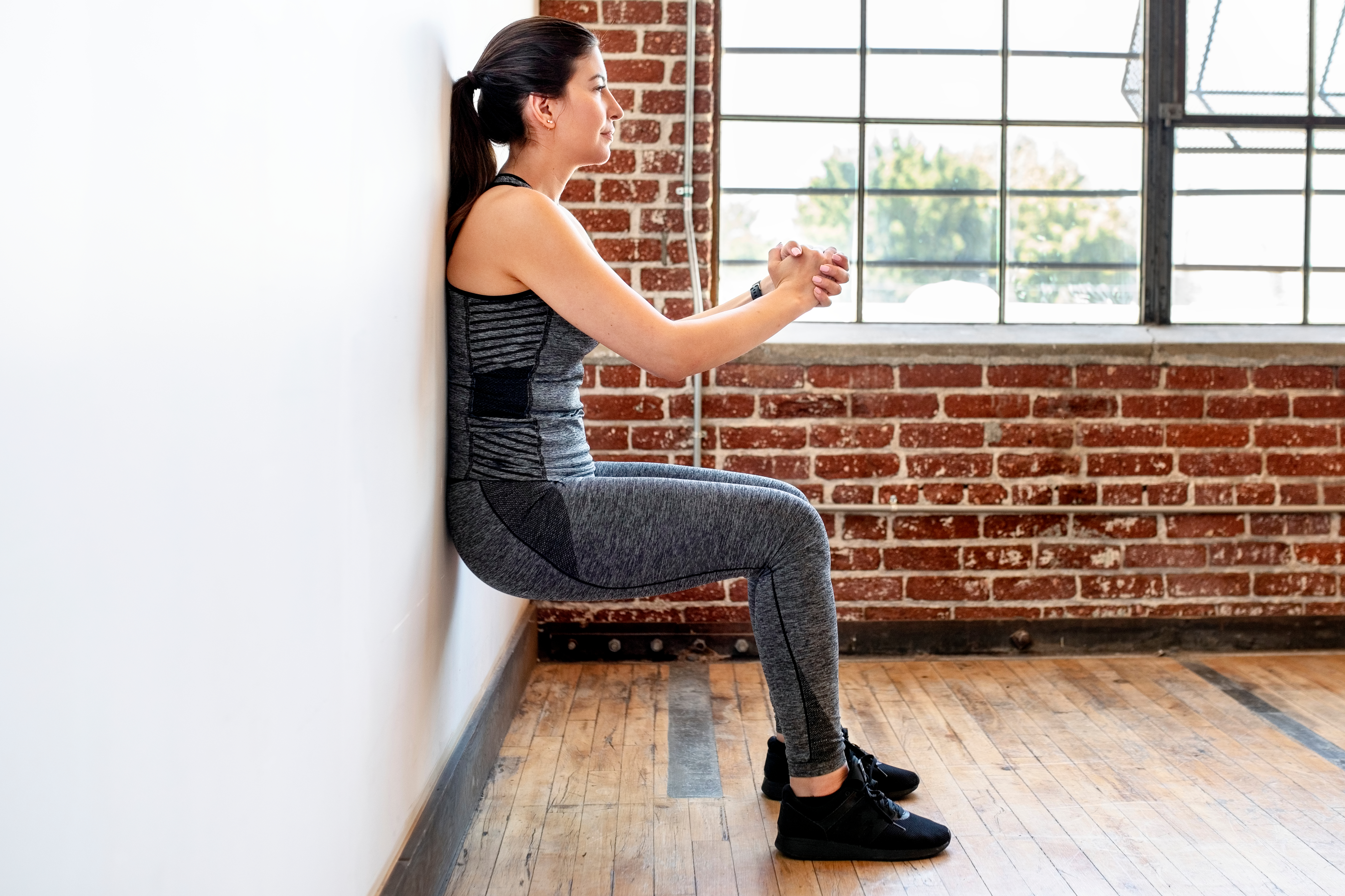 A woman performs a wall sit exercise in a room with exposed brickwork and wooden flooring. Her back is against the wall, knees bent, hands clasped in front of her.