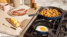 Two blue cast iron skillets on a gas stove, one with fried potato cubes, one with a fried egg. A stick of butter and some fried bacon on the counter beside them. 