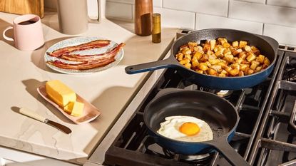 Two blue cast iron skillets on a gas stove, one with fried potato cubes, one with a fried egg. A stick of butter and some fried bacon on the counter beside them. 