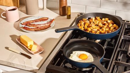 Two blue cast iron skillets on a gas stove, one with fried potato cubes, one with a fried egg. A stick of butter and some fried bacon on the counter beside them. 