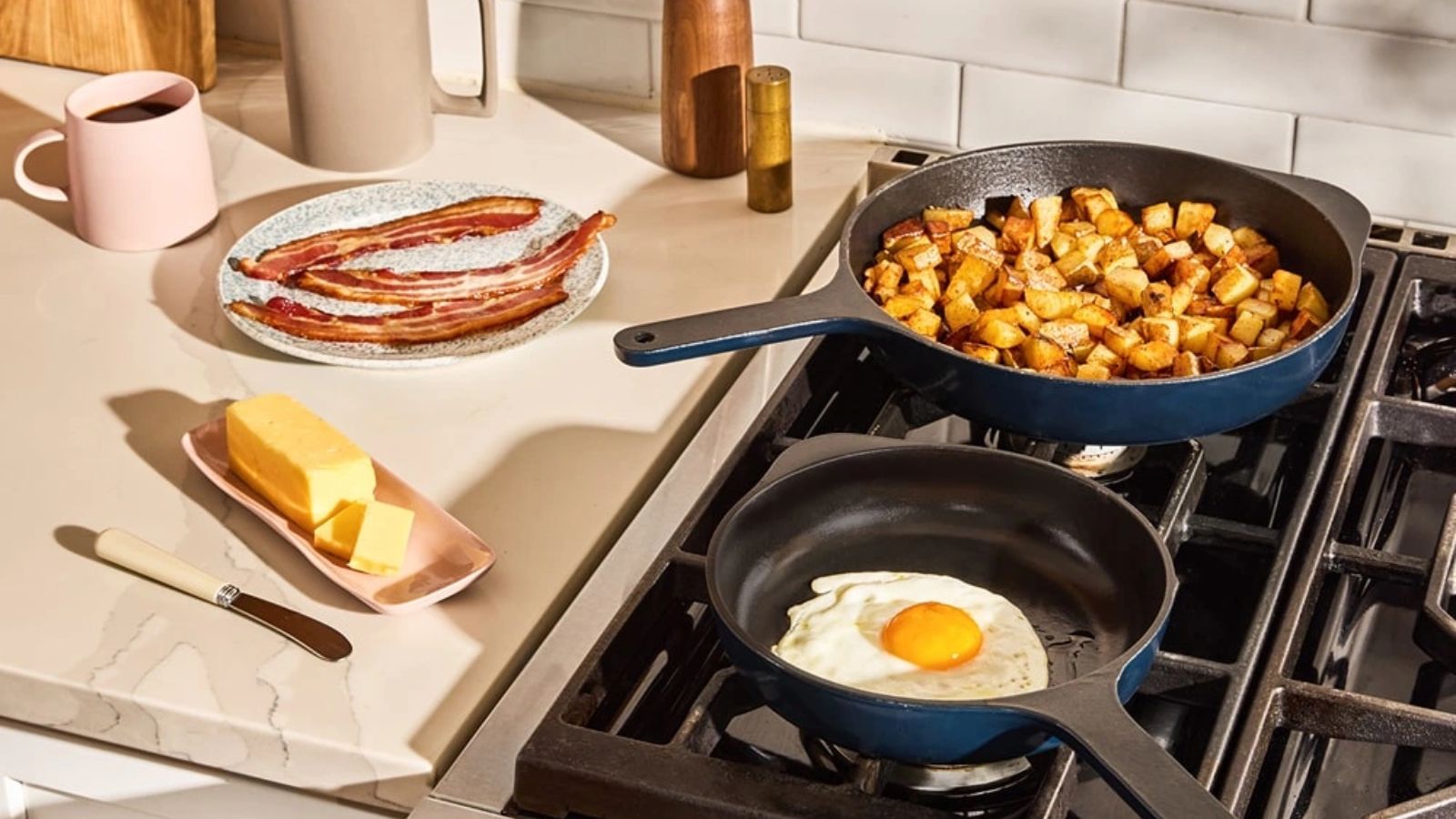 Two blue cast iron skillets on a gas stove, one with fried potato cubes, one with a fried egg. A stick of butter and some fried bacon on the counter beside them. 