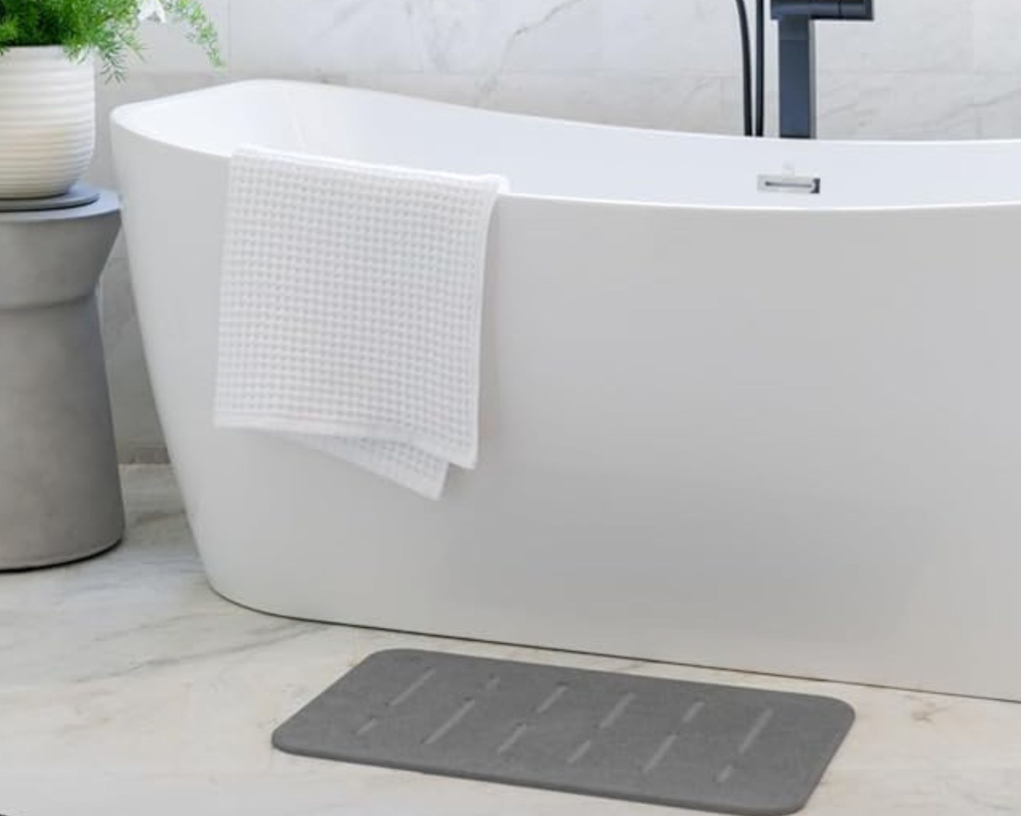 Gray stone bath mat in front of a freestanding white bathtub, and on marble veined floors.
