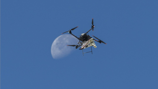 a quadcopter drone carries a wedge-shaped model aircraft in front of a large moon seen in the background