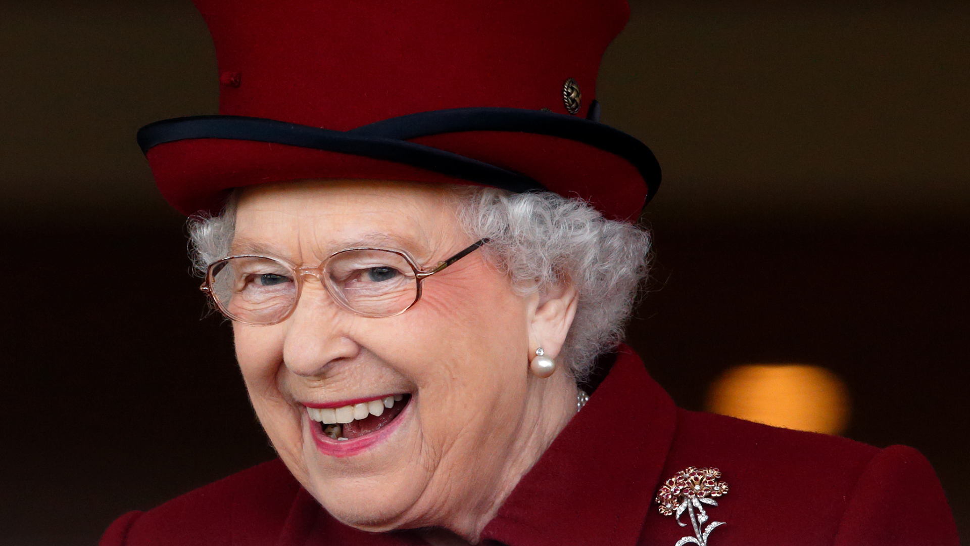 A headshot of Queen Elizabeth wearing a burgundy hat and coat while laughing