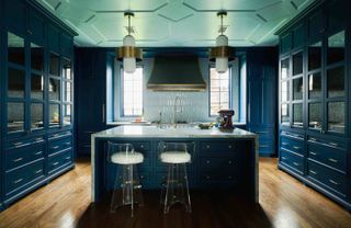 A kitchen island in a dark blue kitchen with marble tops and wood flooring. Two bar stools are placed near the Island.