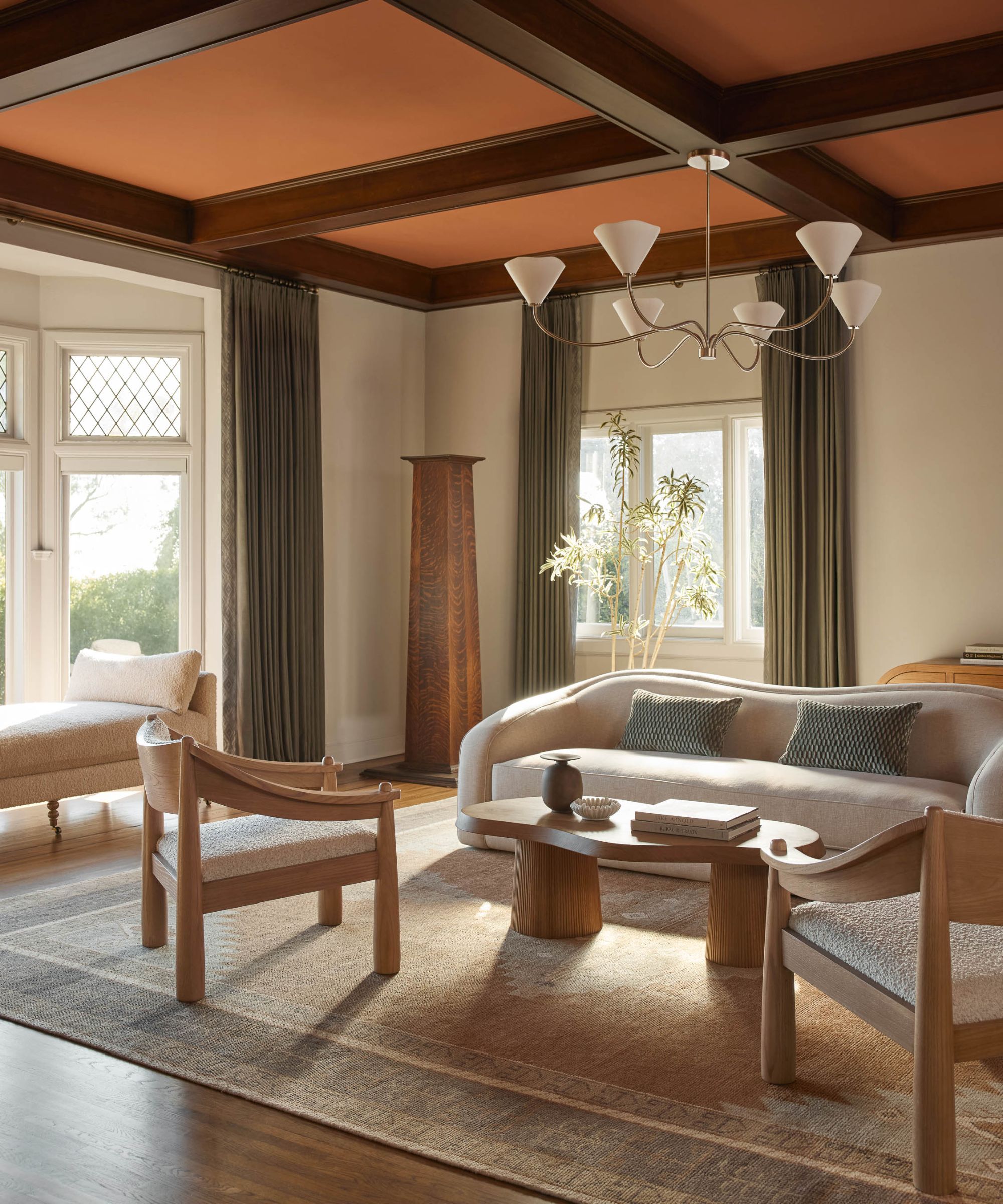 neutral living room with curved boucle sofa, oak coffee table, accent chairs, a large silver chandelier and a beamed wooden ceiling painted orange