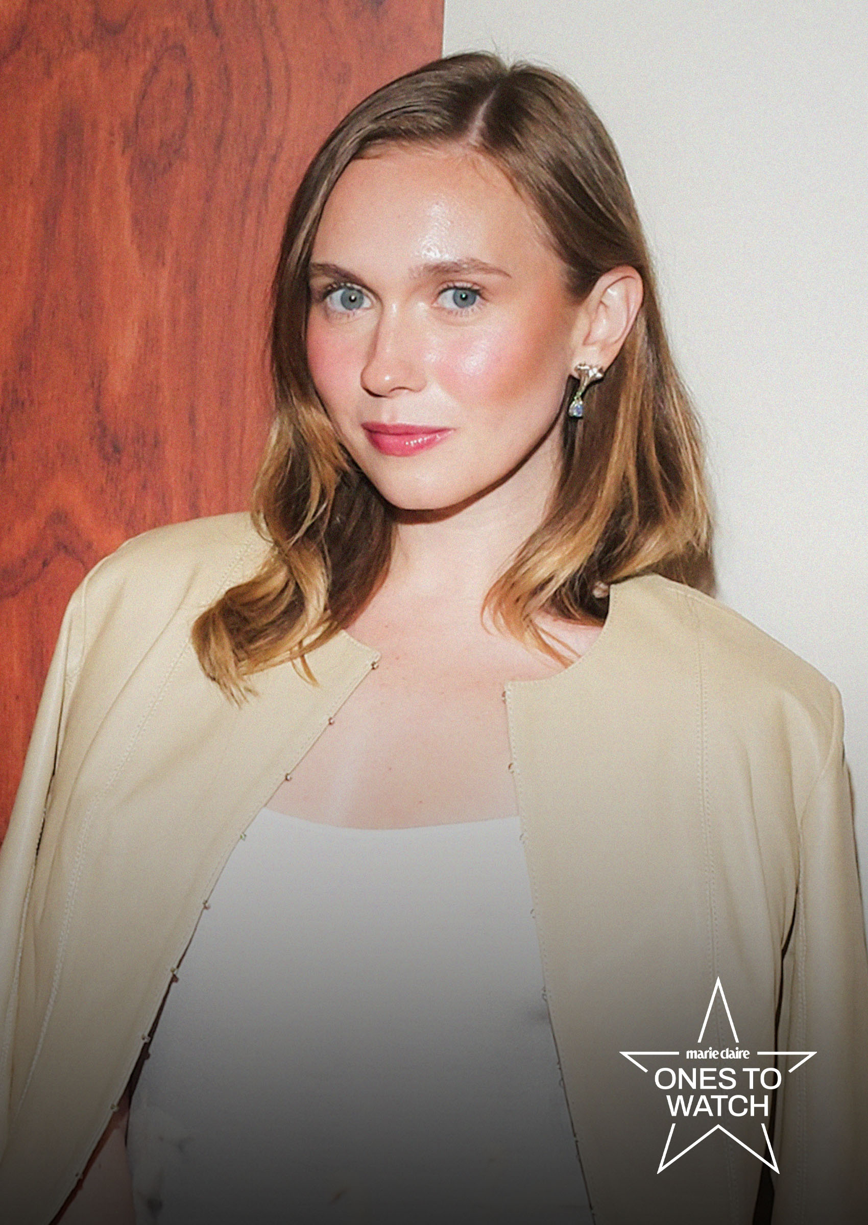 comedian veronika slowikowska wears a tan leather jacket and white dress posing against a wood paneled wall