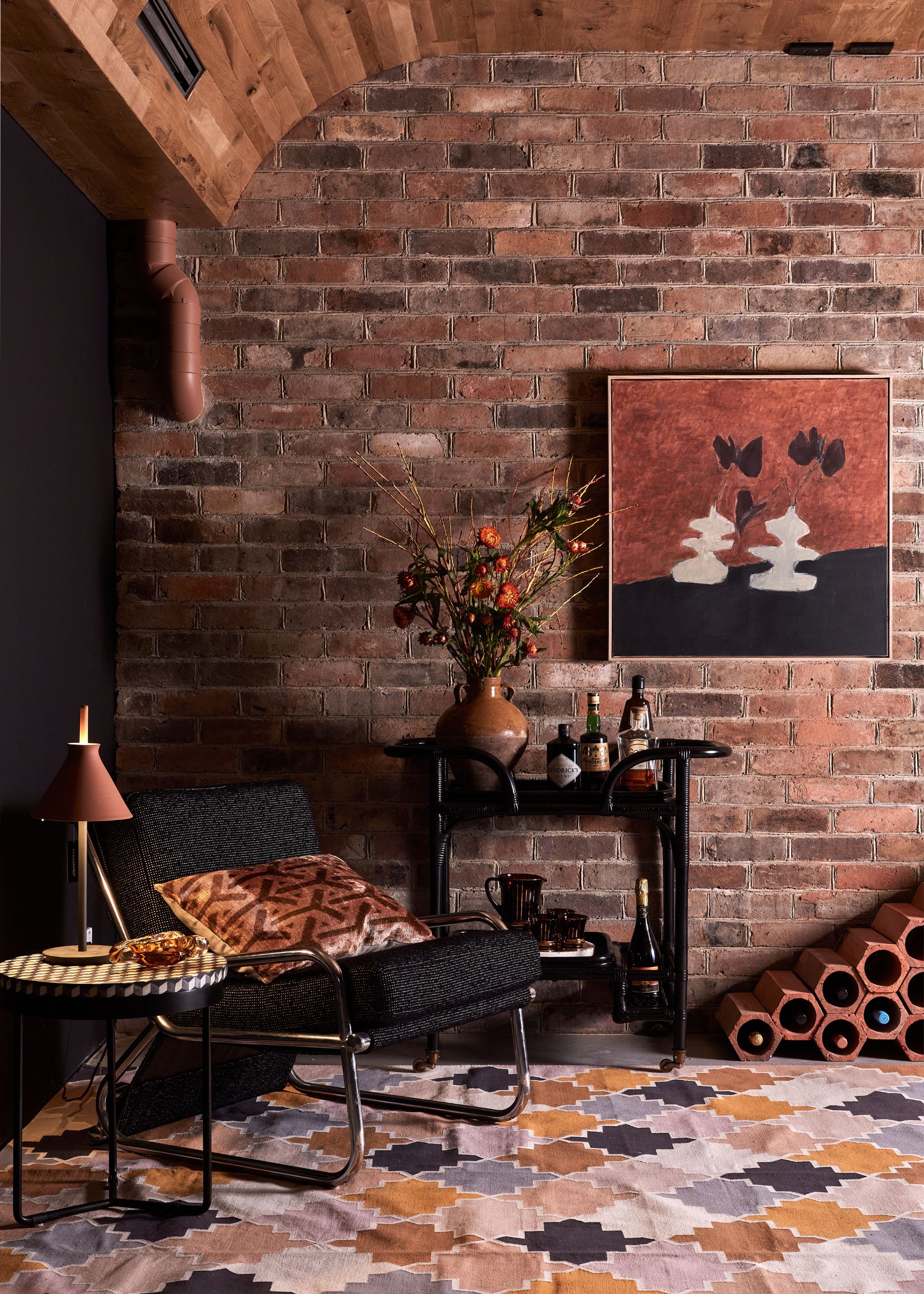A living room with a framed painting, a brick wine rack, a black sofa chair, a side table, a vase of flowers, and a rust table lamp