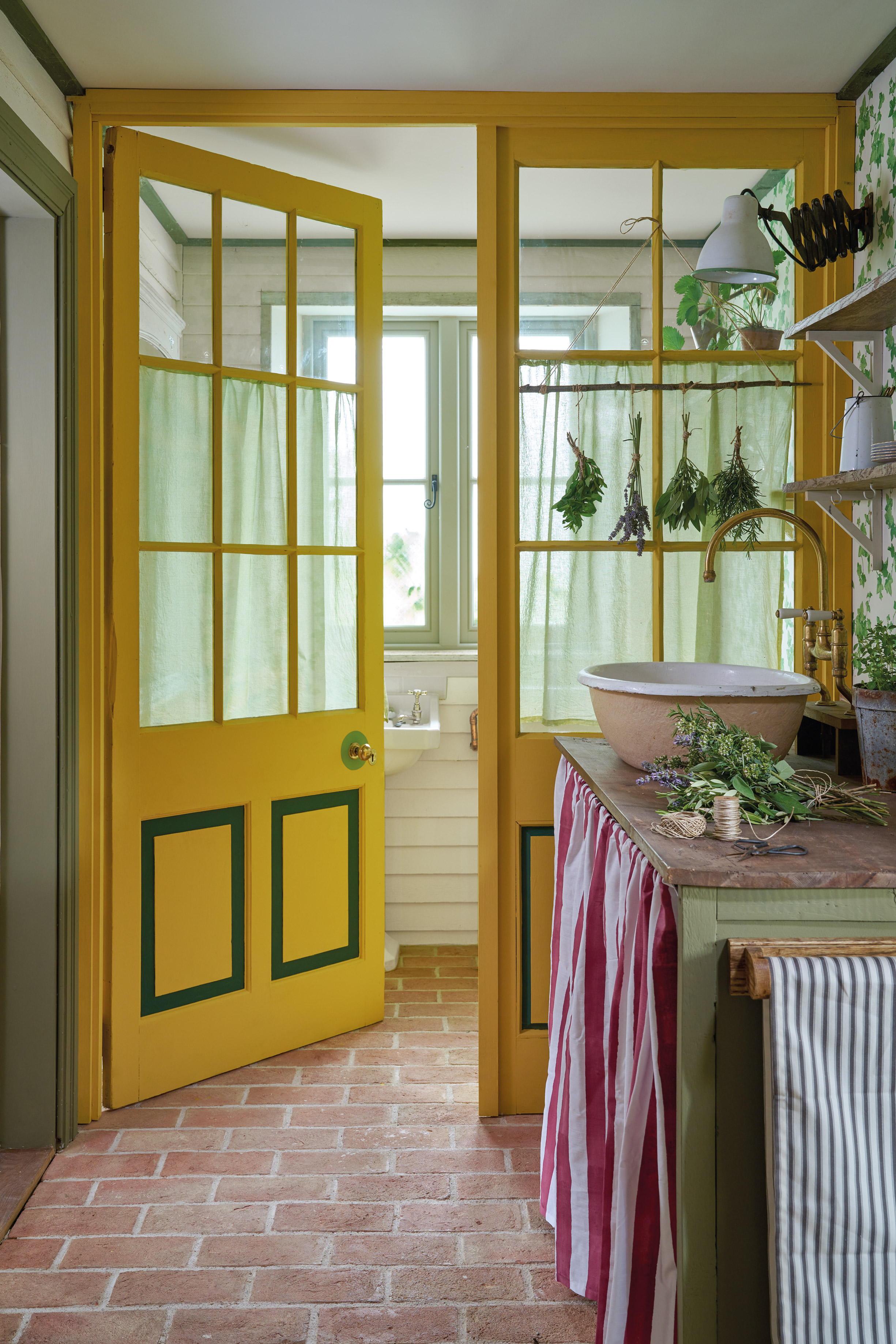 Vibrant utility room with rustic red brick flooring, bright yellow framed doors with green trim, and soft green curtains. A farmhouse-style sink and wooden counter with a bowl and herbs evoke a traditional country kitchen feel.