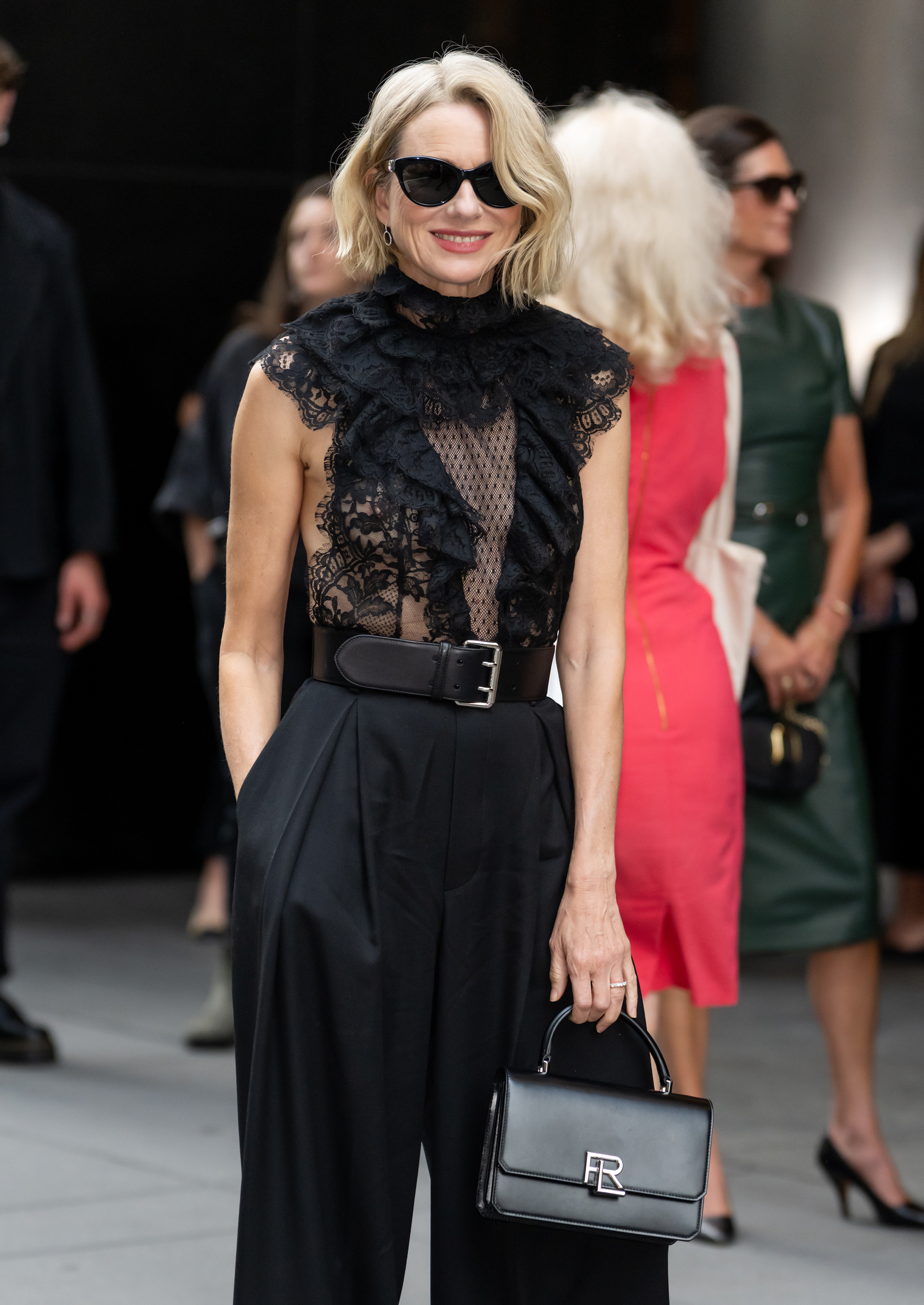 A woman with wavy, short, blonde hair wearing a lace top and black pants smiling at the camera. She is holding a black handbag.