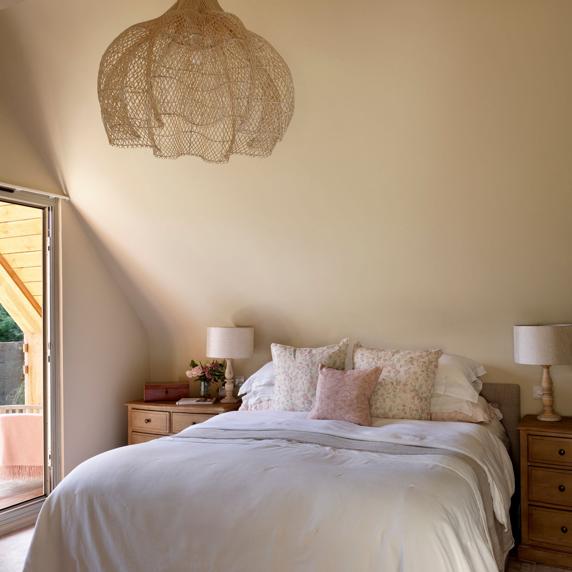 Beige bedroom with double bed and floral cushions, wooden bedside tables with white lamps and a large pendant light