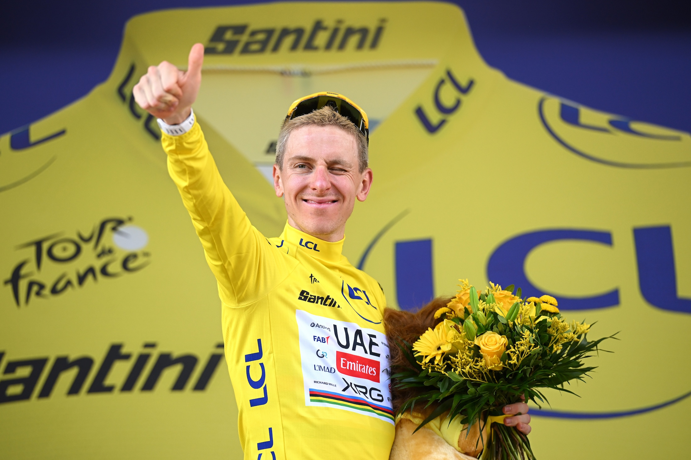 Pogacar in the yellow jersey holding flowers gives a thumbs up on the podium