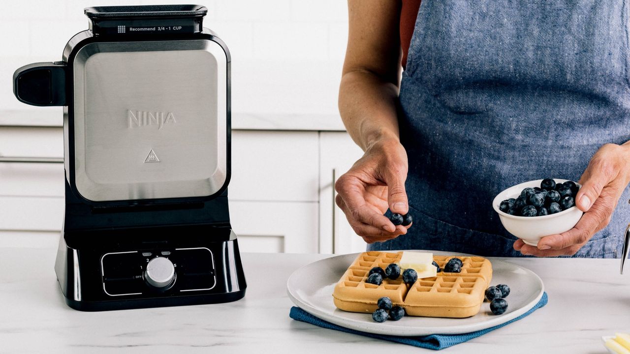 Person adding blueberries to waffle beside Ninja Belgian Waffle Maker
