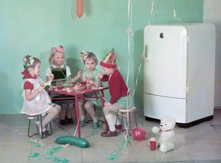 Four children in party hats sitting at pink table in from of white refrigerator